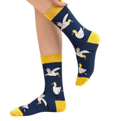 A pair of navy blue crew socks with yellow cuffs, heels, and toes, featuring a pattern of gannets in flight, shown on a person's feet against a white background.
