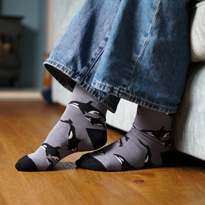 Person wearing gray socks with orca patterns and blue jeans on a wooden floor.